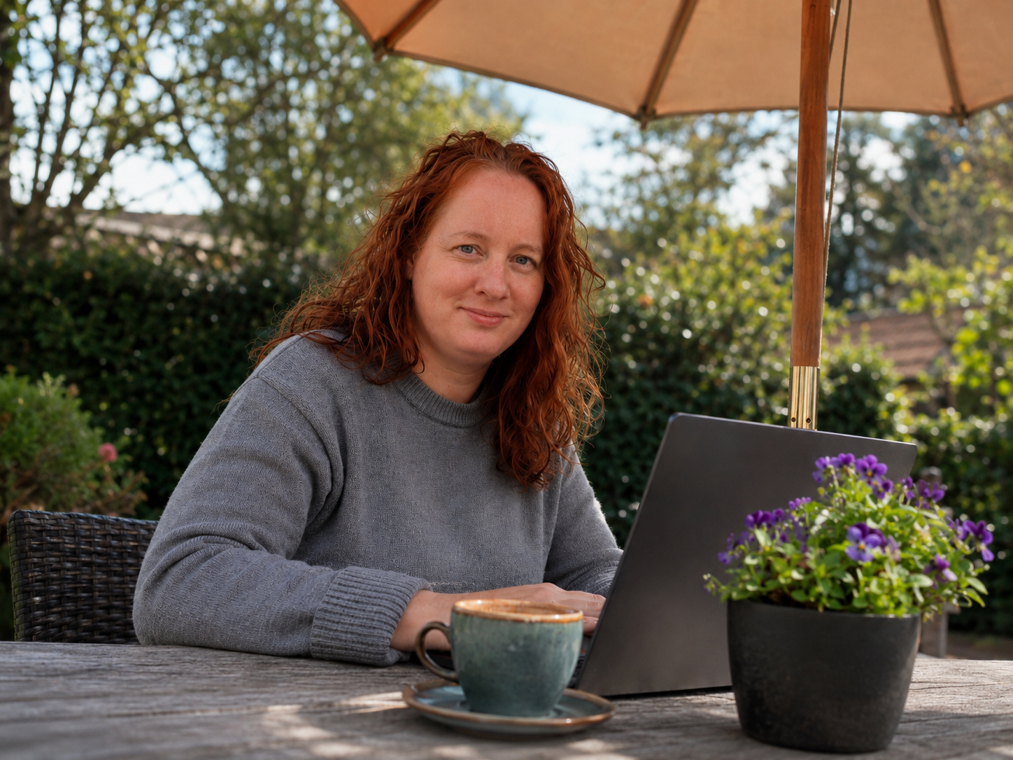 Vrouw met rood haar werkt op een openlucht terras onder een paraplu, met een laptop, een kopje koffie en een bloempot met paarse bloemen op tafel.