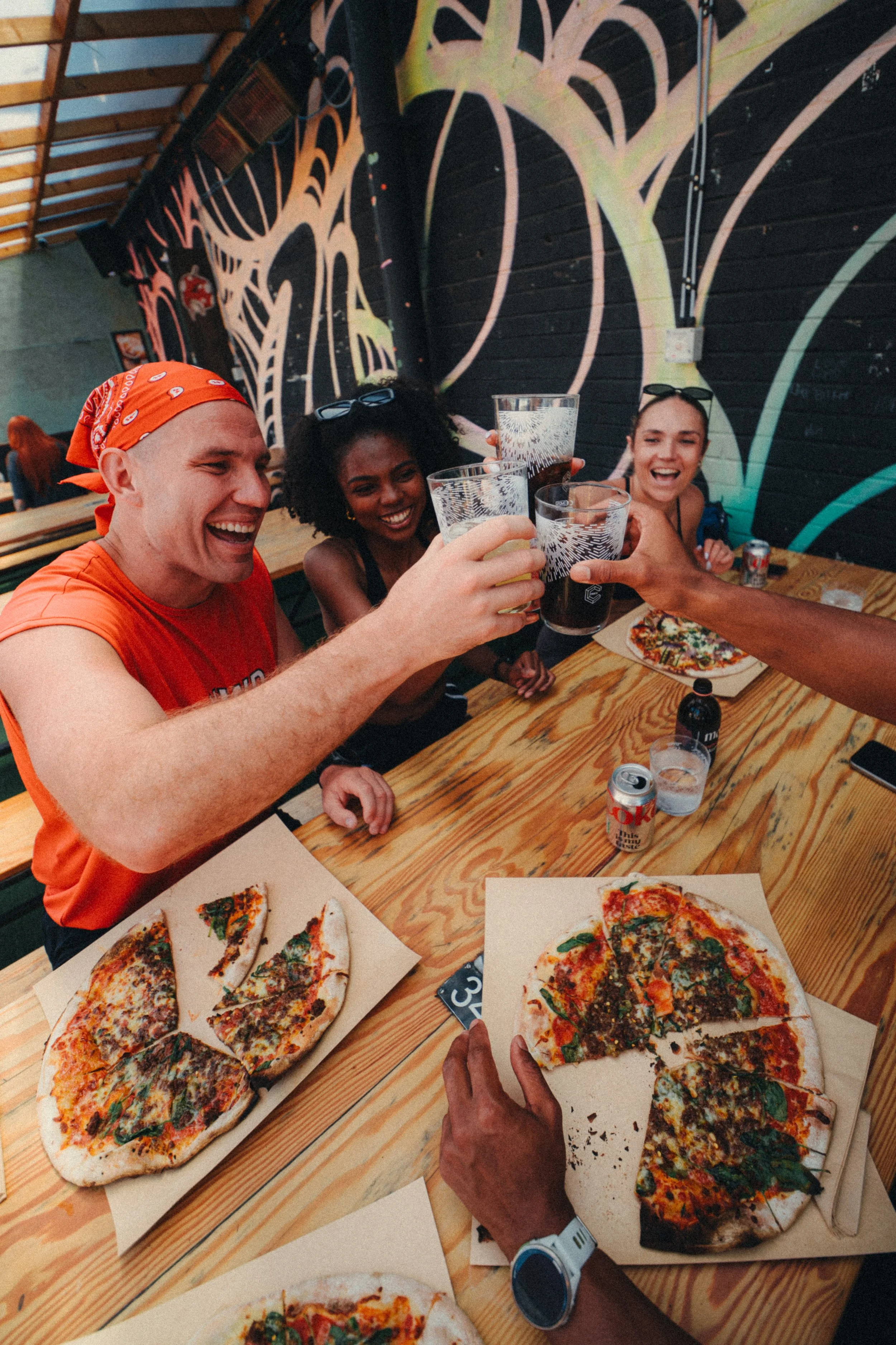 Group of friends enjoying drinks and pizza at a wooden table in a restaurant or bar with abstract black and yellow wall art.