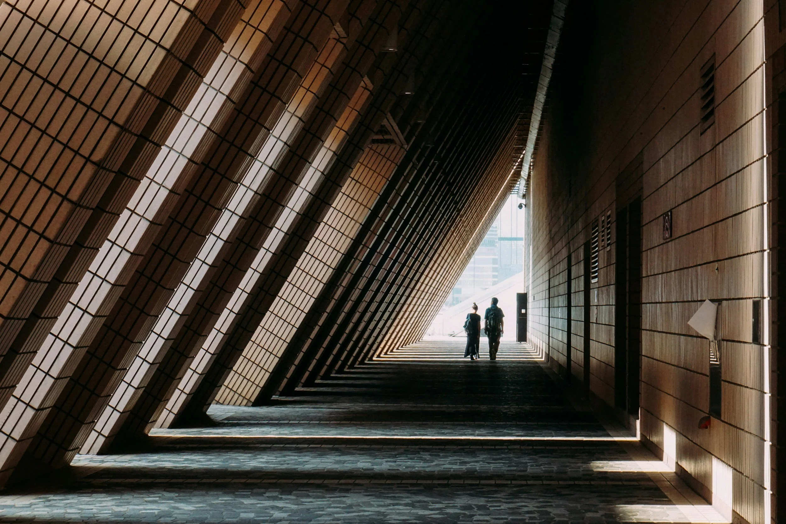 Two people walk through a modern architectural corridor with a wooden slatted ceiling and walls, geometric patterned floor, and natural light at the end of the passage.
