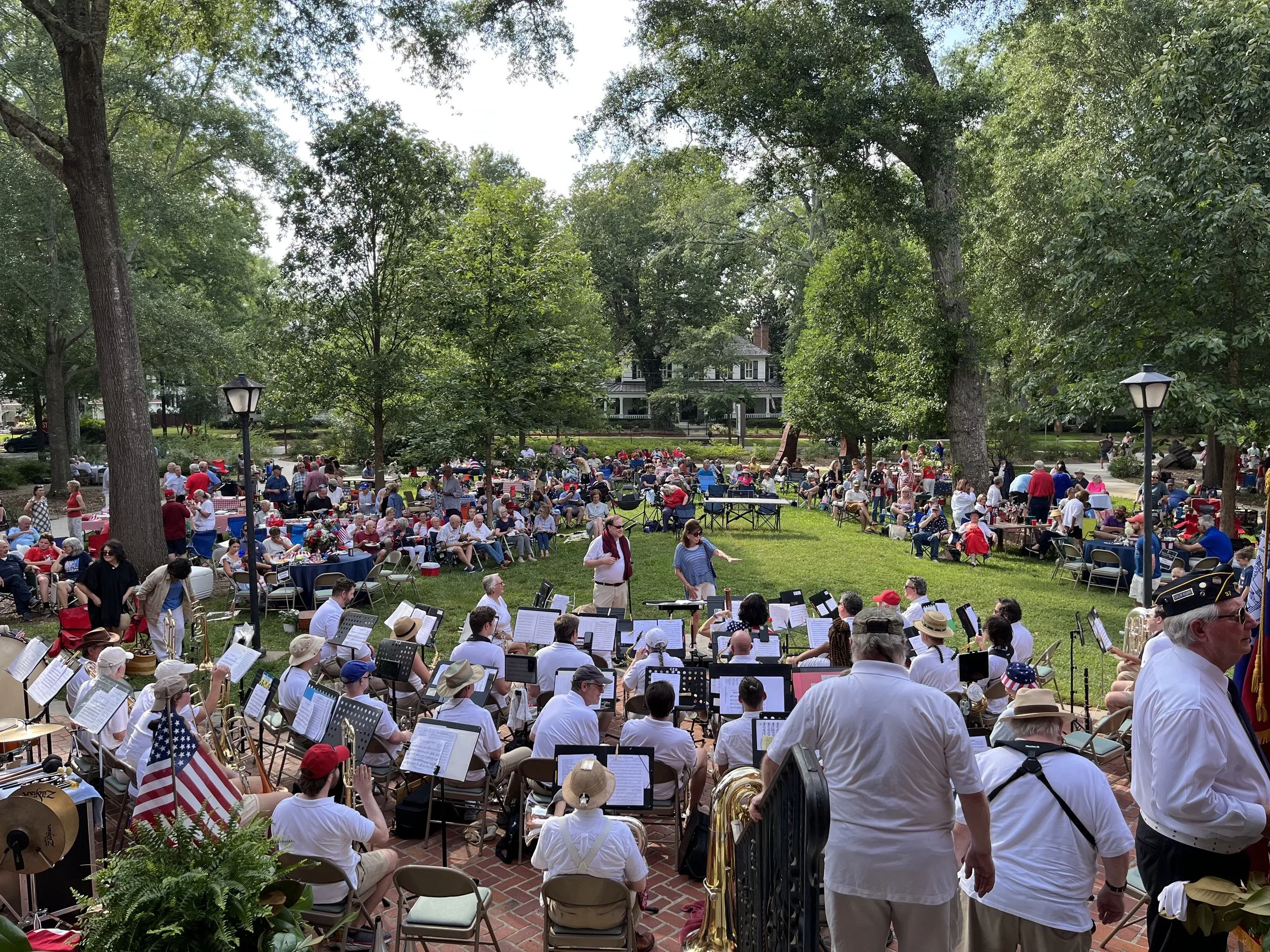Large outdoor gathering with a community event on a grassy park, featuring an orchestra playing music, surrounded by people sitting and standing, some wearing patriotic hats and accessories, with trees and houses in the background.