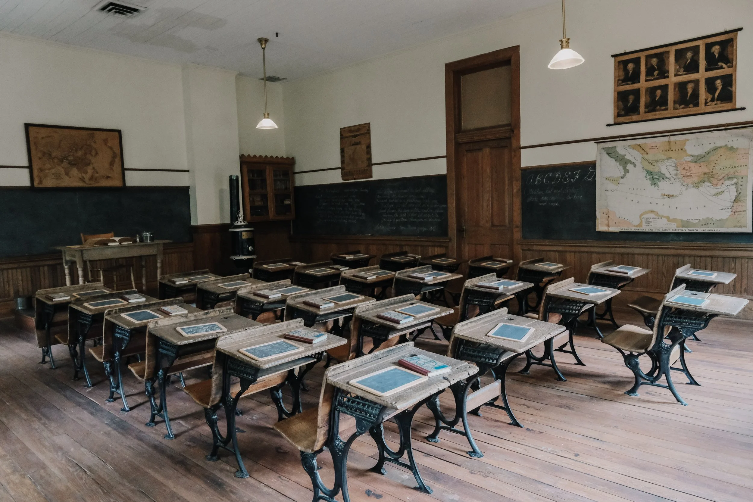An empty classroom with wooden desks and chairs, a blackboard with writing, maps and paintings on the walls, and pendant lights hanging from the ceiling.