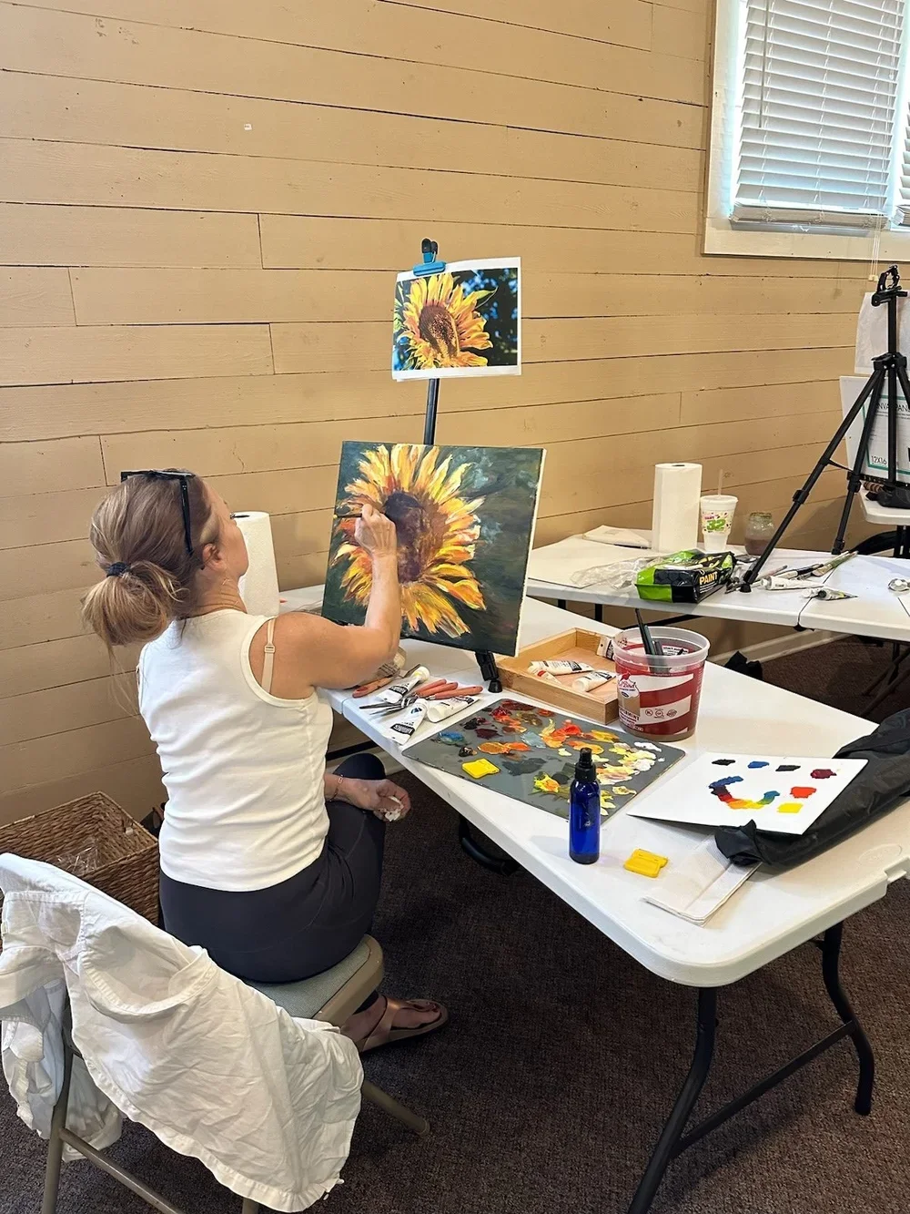 A woman painting a sunflower on a canvas in an art classroom.