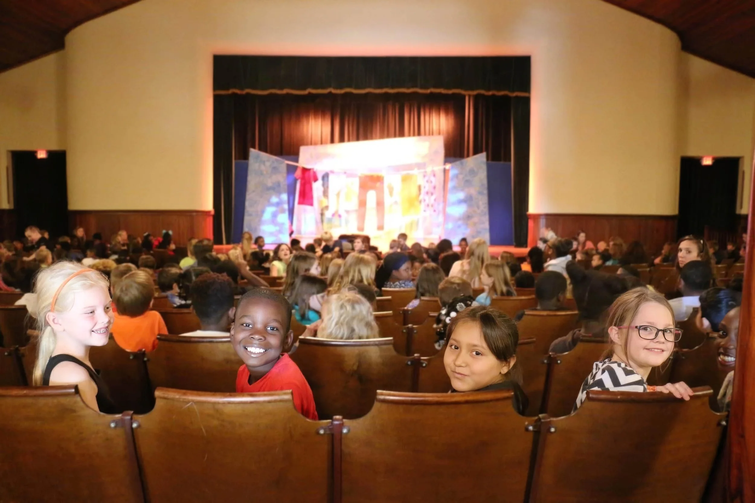 A large auditorium filled with children sitting in rows of wooden chairs, smiling and looking towards the camera. The stage at the front has colorful decorations and a backdrop of dark curtains, with lighting accentuating the stage area.