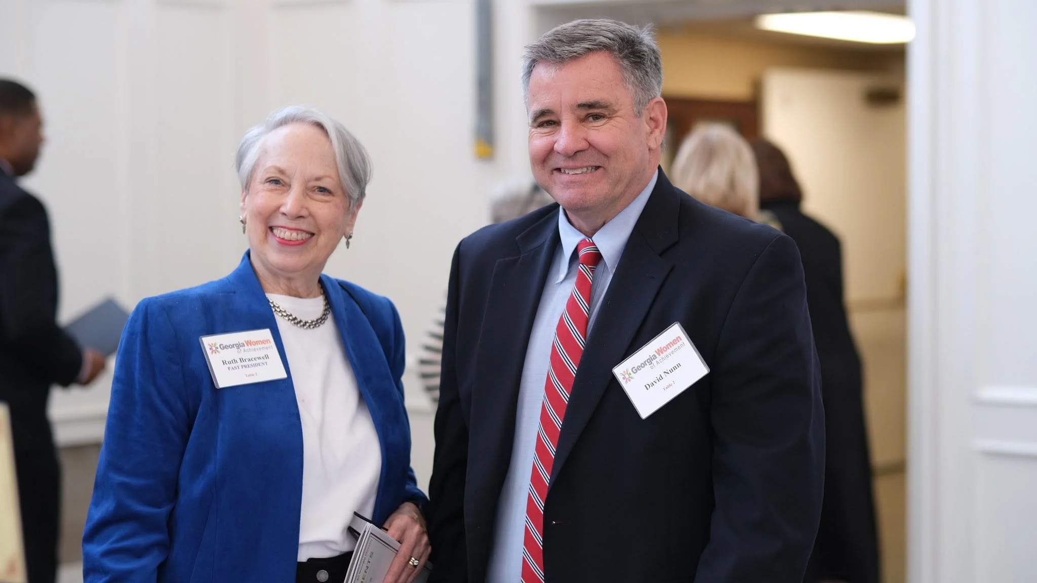 Two people, a woman with short gray hair in a blue blazer and a man with gray hair in a dark suit and red striped tie, smiling and standing close together at a professional event, both wearing name tags that read 'Georgia Women of Achievement.'