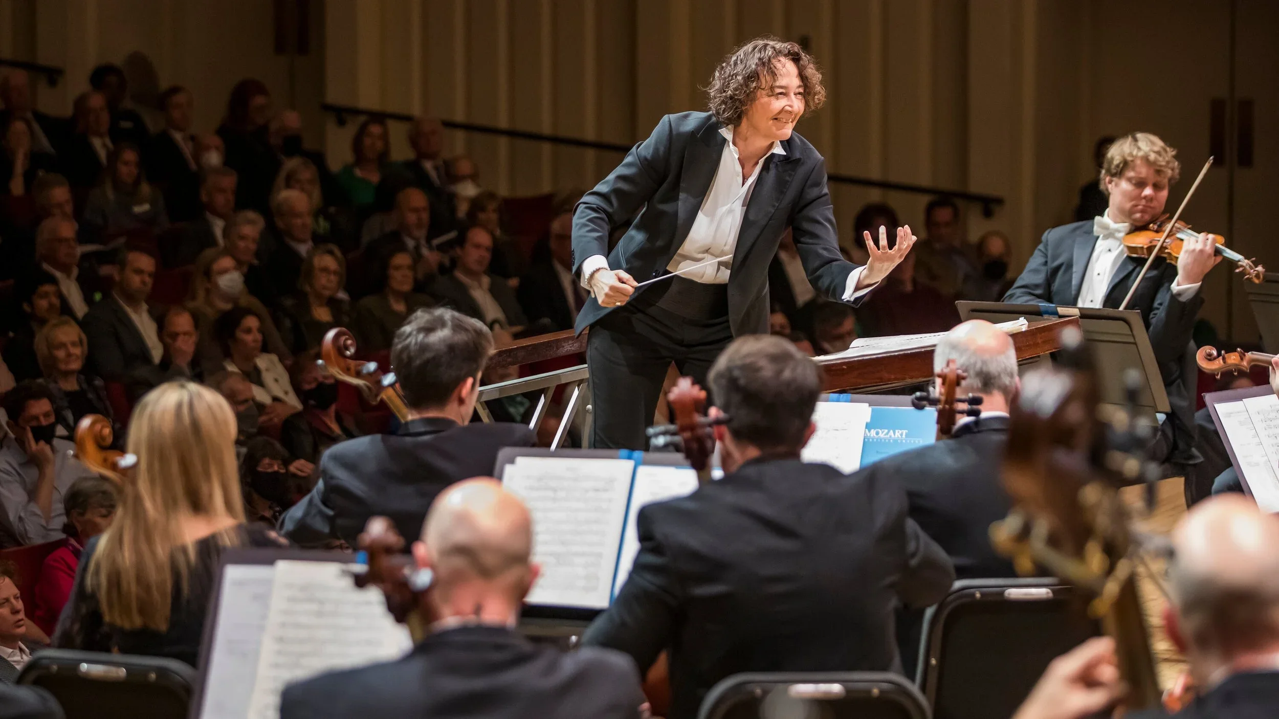 Orchestra performing on stage with a conductor and a violinist, in front of an audience.