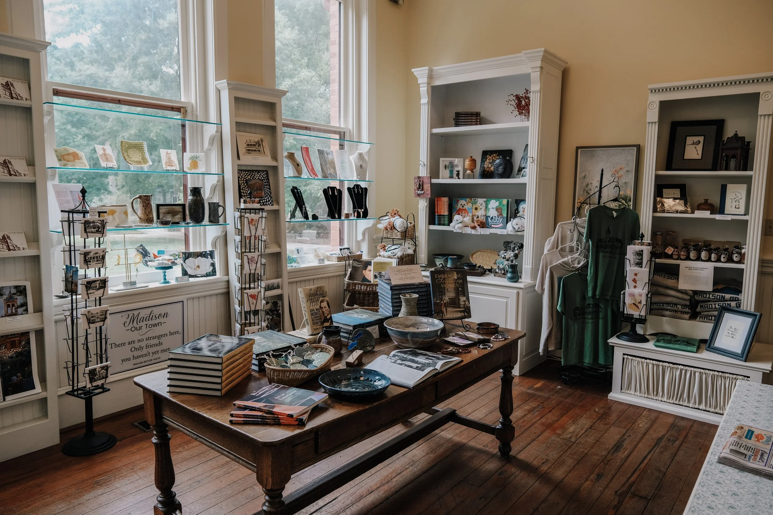 Interior of a gift shop with display shelves holding artwork, jewelry, and small decorative items. A wooden table in the center displays books, pottery, and souvenirs, with large windows letting in natural light.