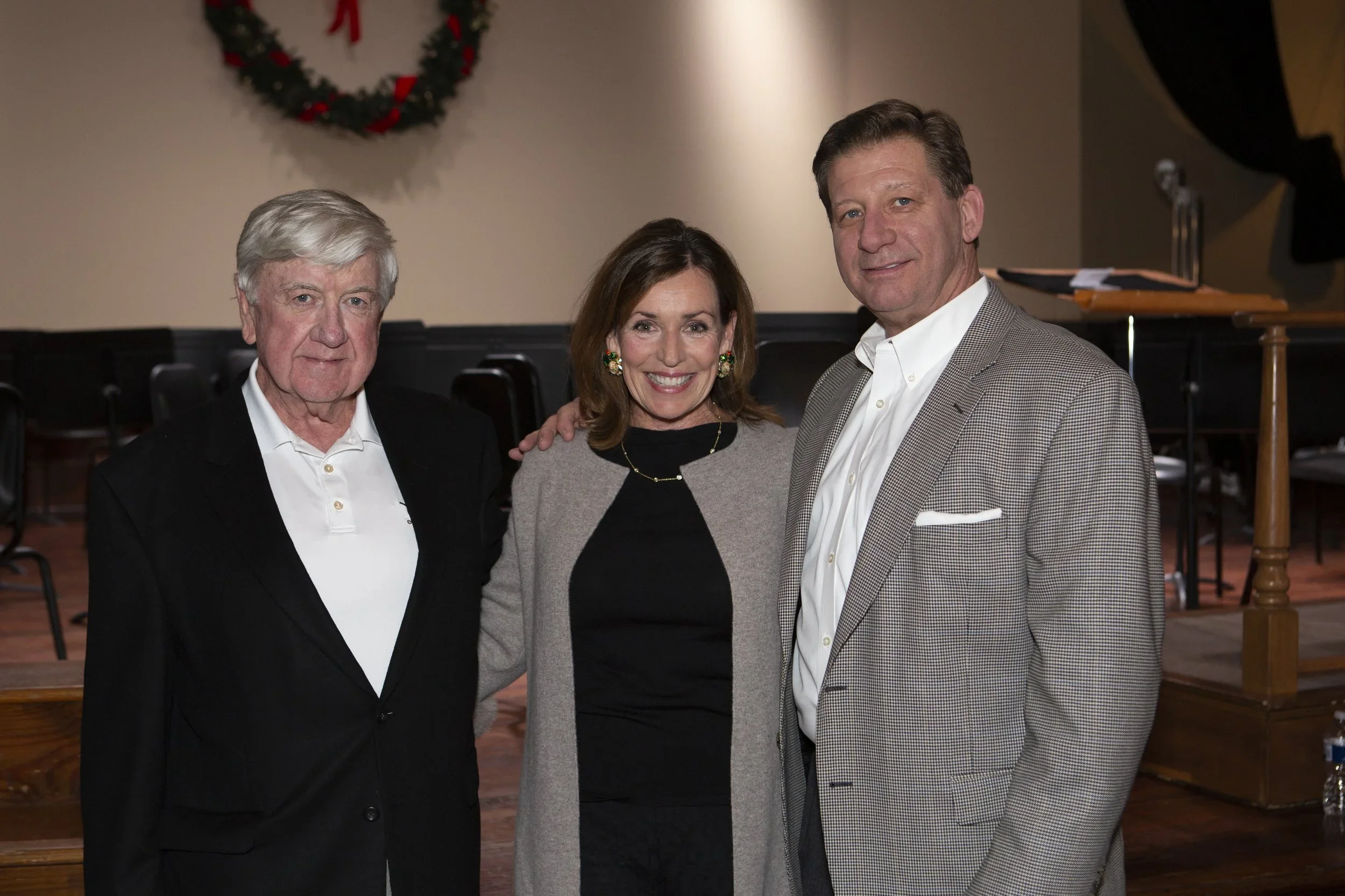 Three smiling middle-aged adults standing together indoors, with a Christmas wreath on the wall behind them, dressed in formal and semi-formal attire.