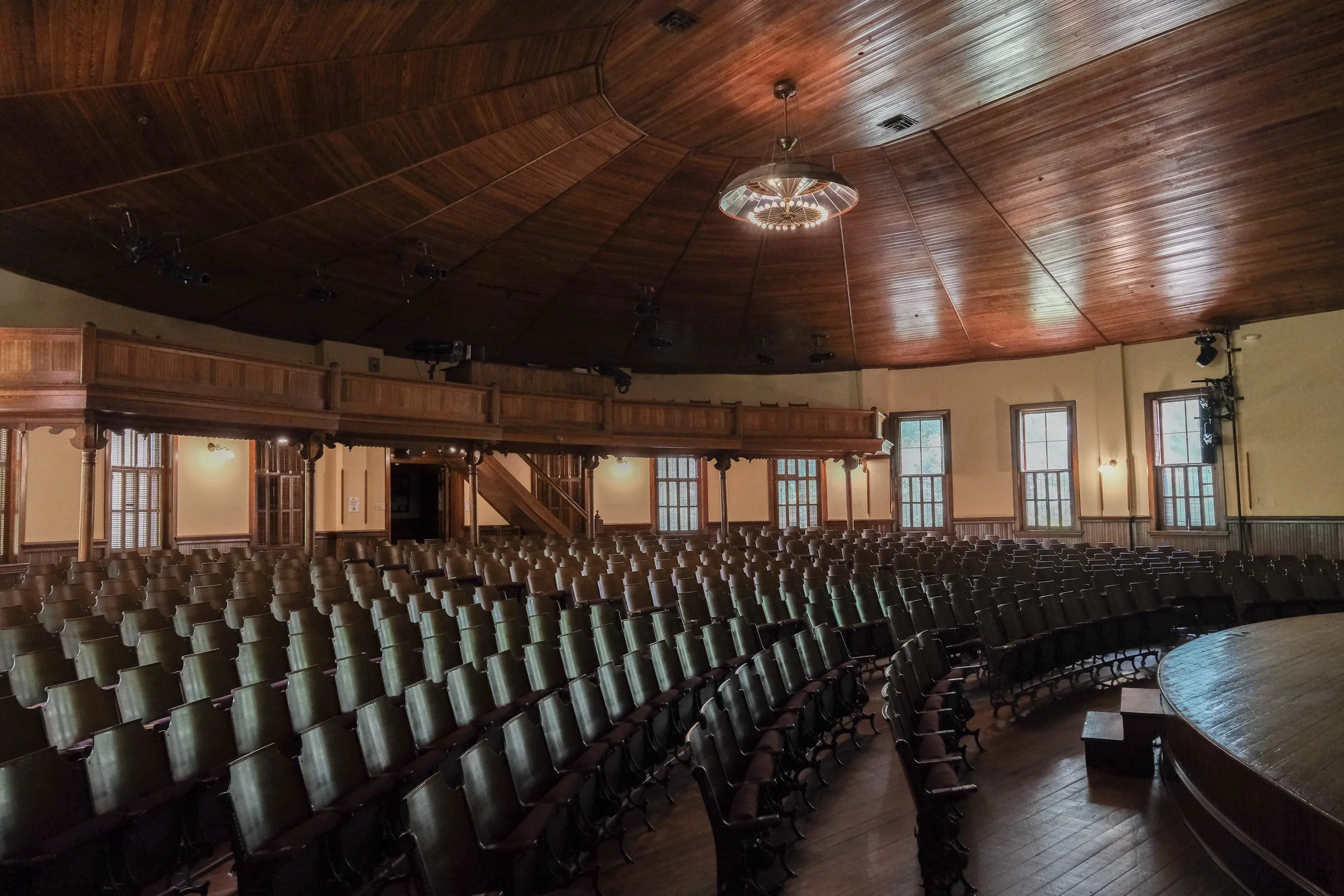 An empty auditorium with rows of chairs, a wooden balcony, a wood-paneled ceiling, large windows, and a stage area.