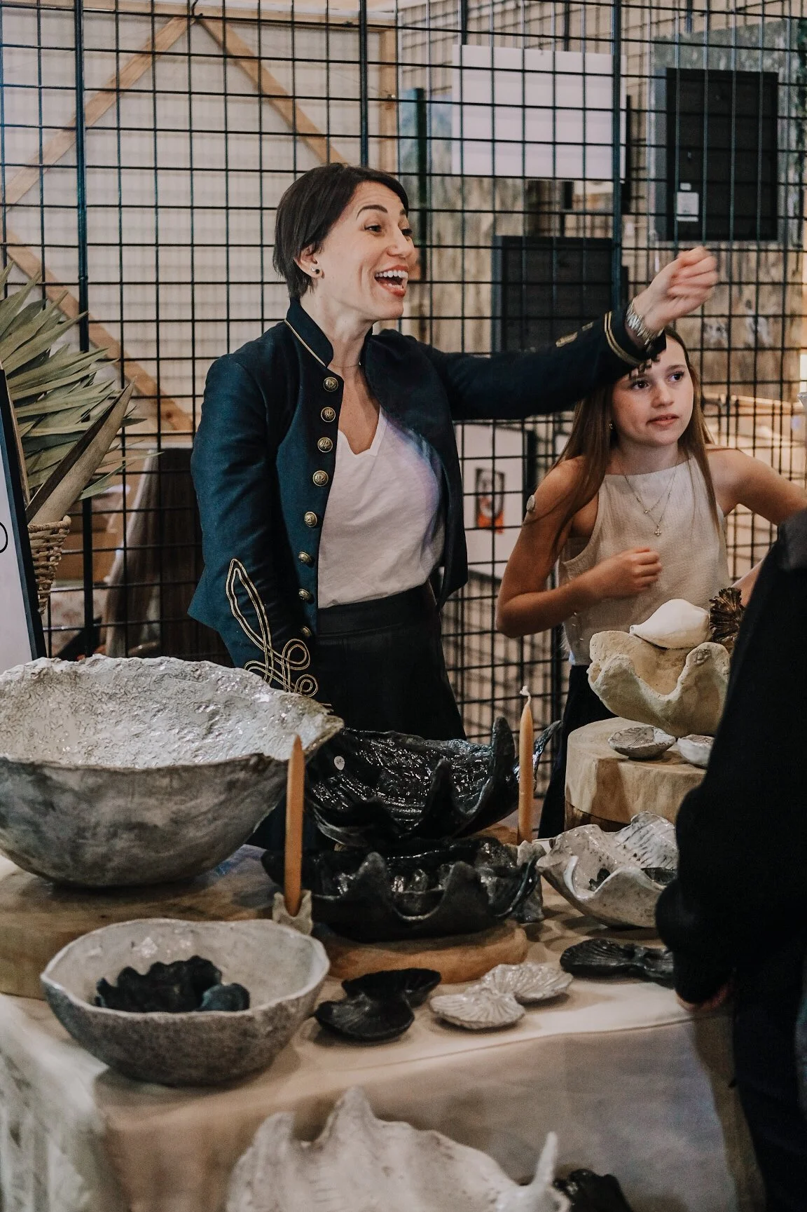 A woman with short dark hair wearing a black and gold jacket talking to a girl with long brown hair at a display of various ceramic bowls and sculptures, in an indoor setting with a metal grid wall in the background.