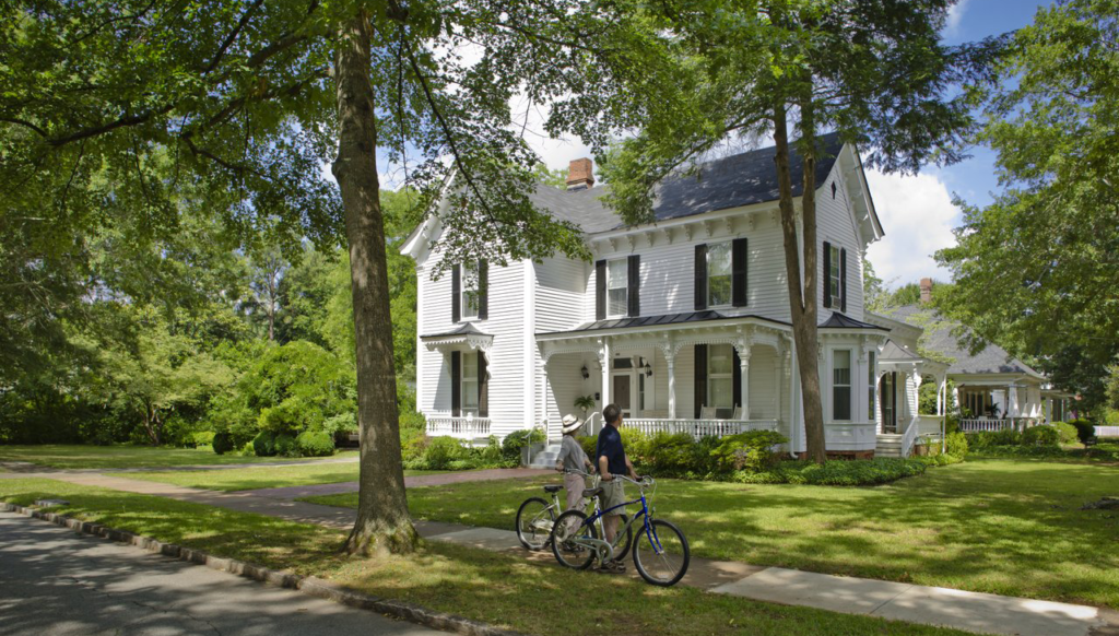 Two people with bicycles walking past a large white Victorian house with a porch, surrounded by green trees and lawn on a sunny day.