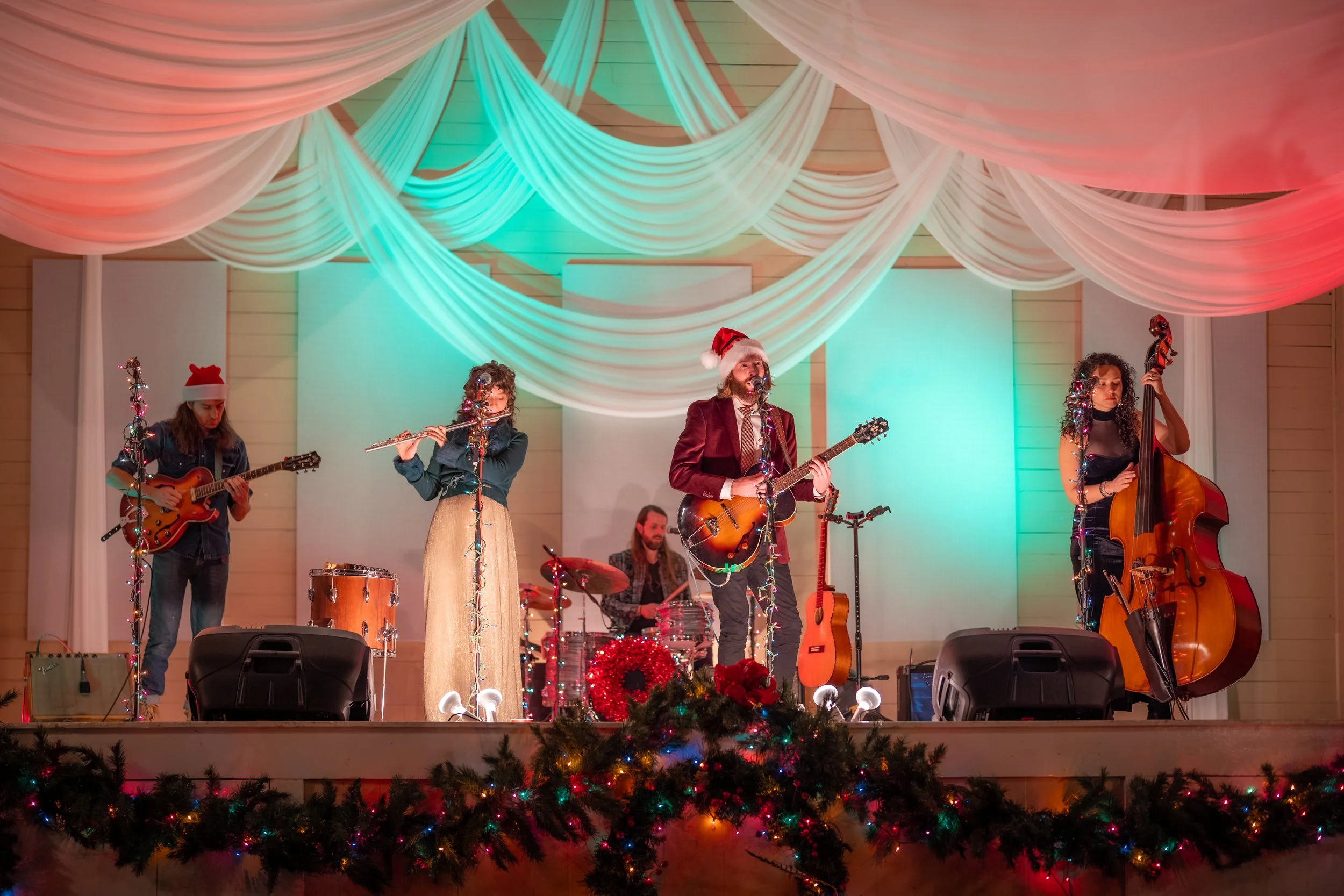 A band performing on stage decorated for Christmas, with draped white fabric, colorful lights, and Christmas wreaths and garlands in the foreground. The band members are wearing festive accessories like Santa hats, and they are playing guitars, a drum, a ukulele, a flute, and a double bass.