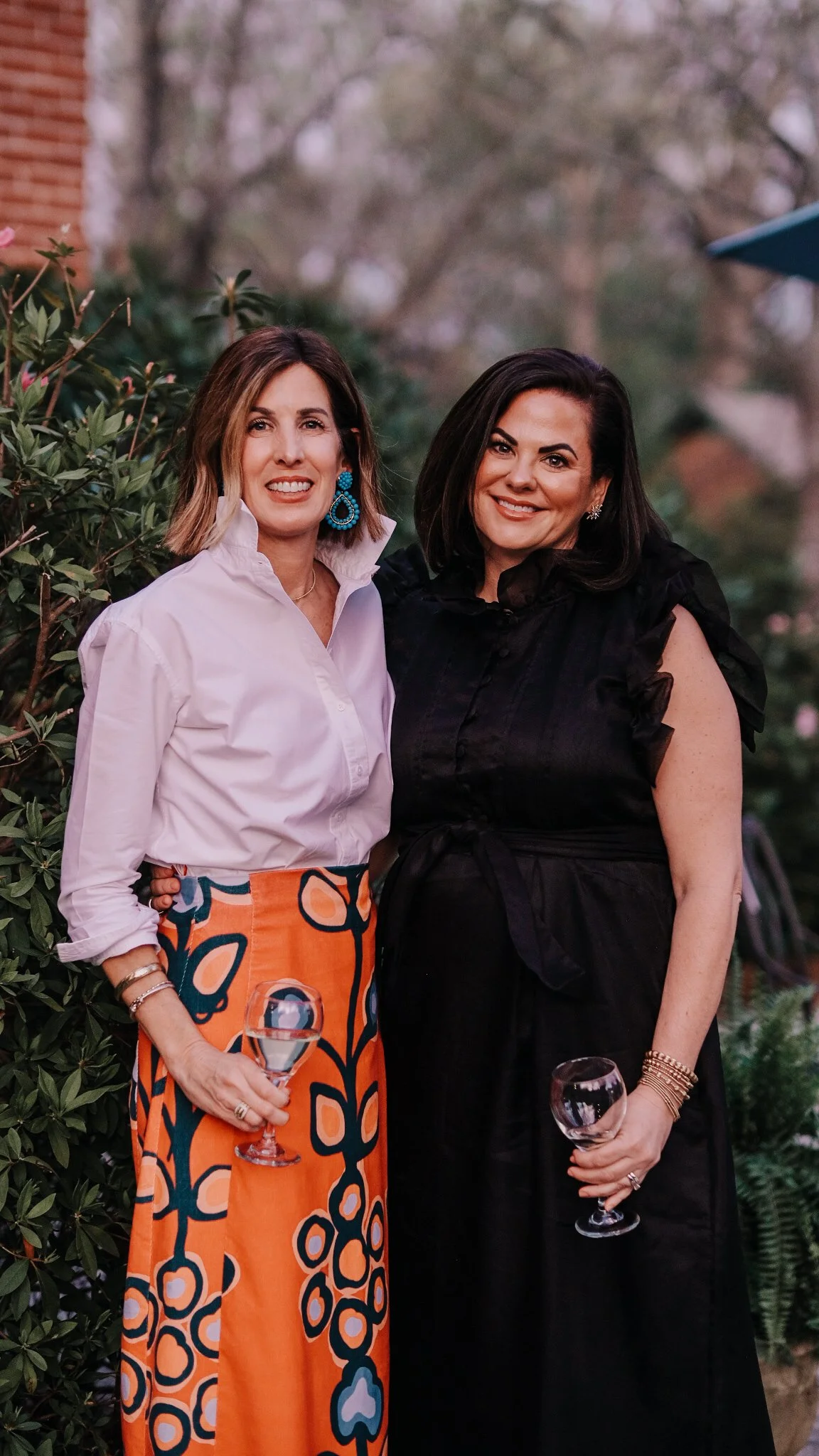 Two women standing outdoors, holding wine glasses and smiling, with greenery and trees in the background.