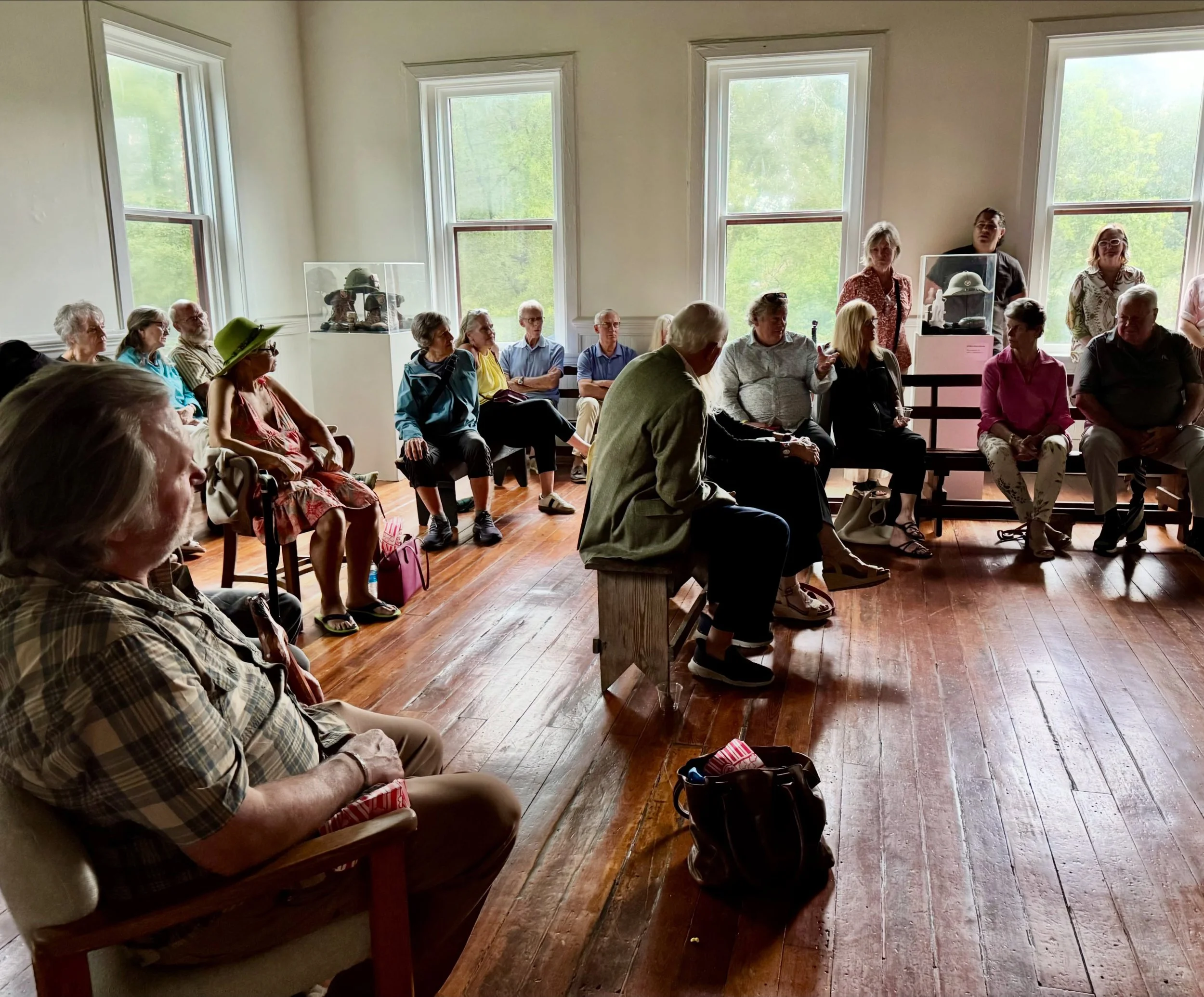 A group of people sitting and standing in a room with wooden floors and large windows, attended a presentation or discussion. Some are engaged, others are listening or looking around.