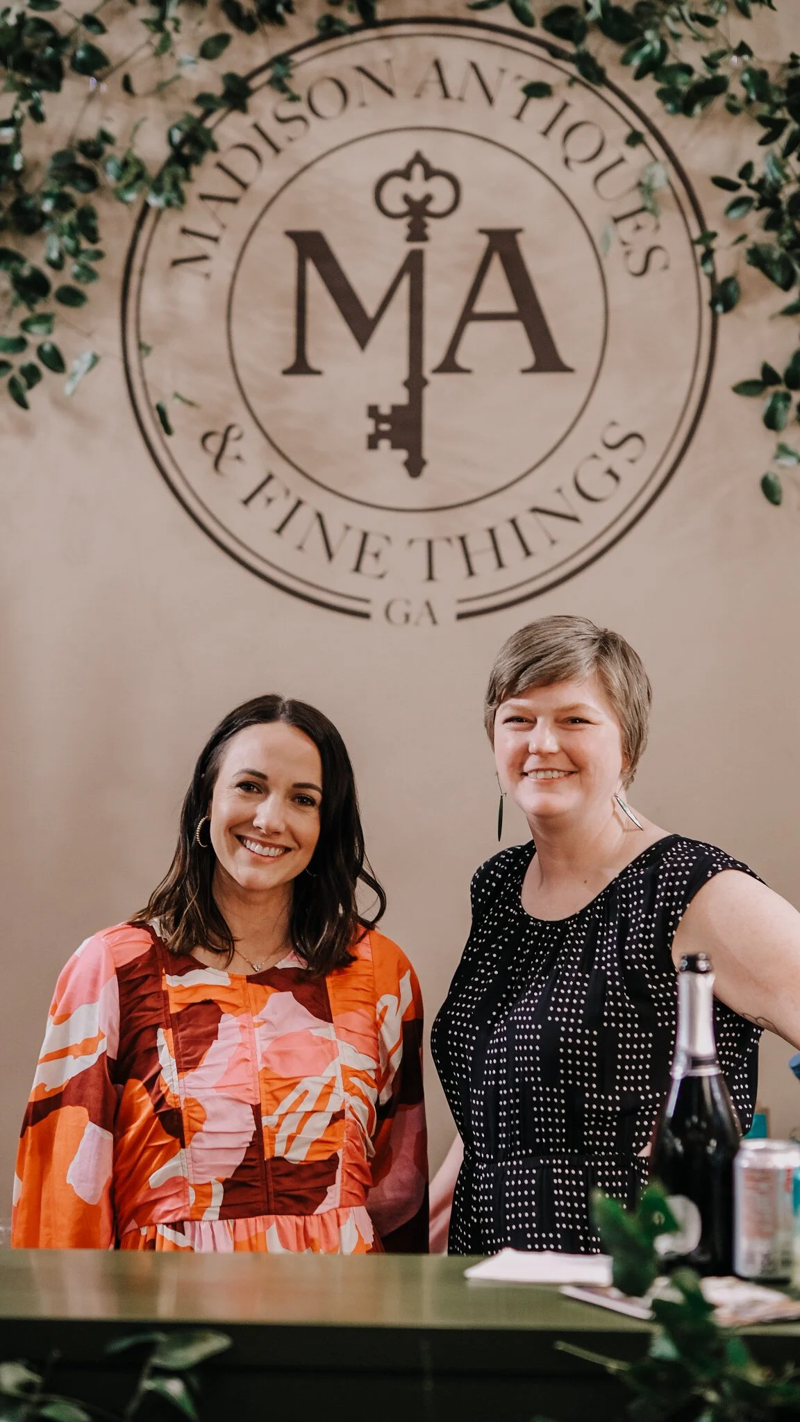 Two women smiling in front of a wall with a circular sign that reads 'Madison Antiques & Fine Things, GA'. The woman on the left has long dark hair and is wearing a colorful patterned blouse. The woman on the right has short light brown hair and is wearing a black dress with white polka dots. There are some green plants in the foreground, and bottles and a bottle opener are partially visible on the counter in front of them.