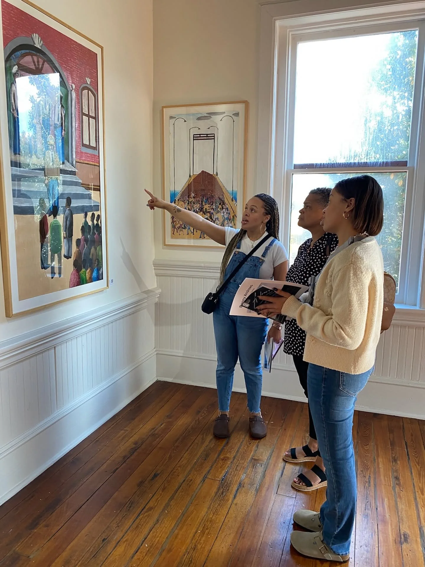 Three women standing in an art gallery, observing a large framed painting on the wall. One woman is pointing at the painting, while the other two look on. The room has wooden floors and large windows.