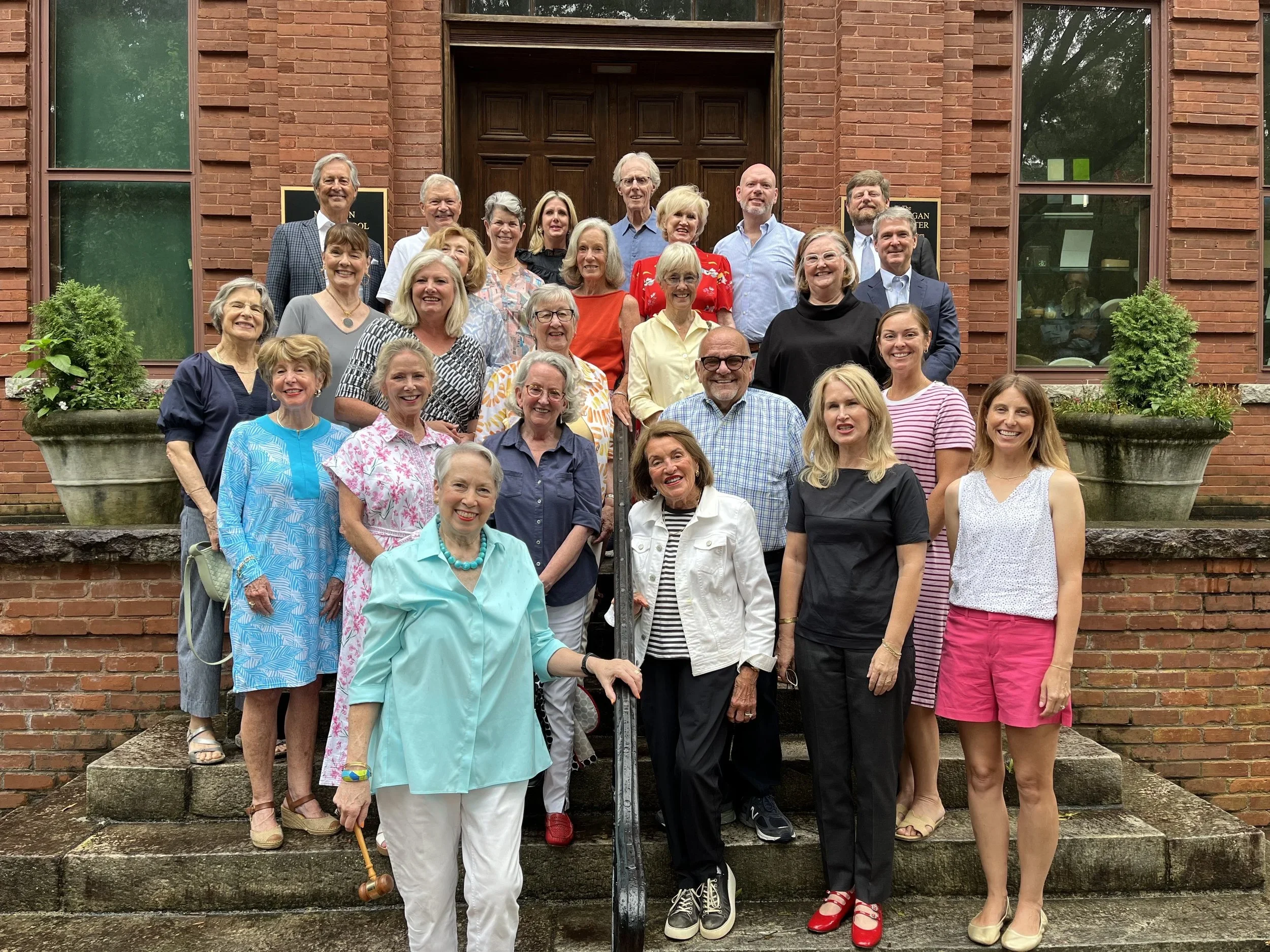A large group of diverse people, mostly women, gathered on the steps outside a brick building with wooden doors, smiling for a group photo in daytime.