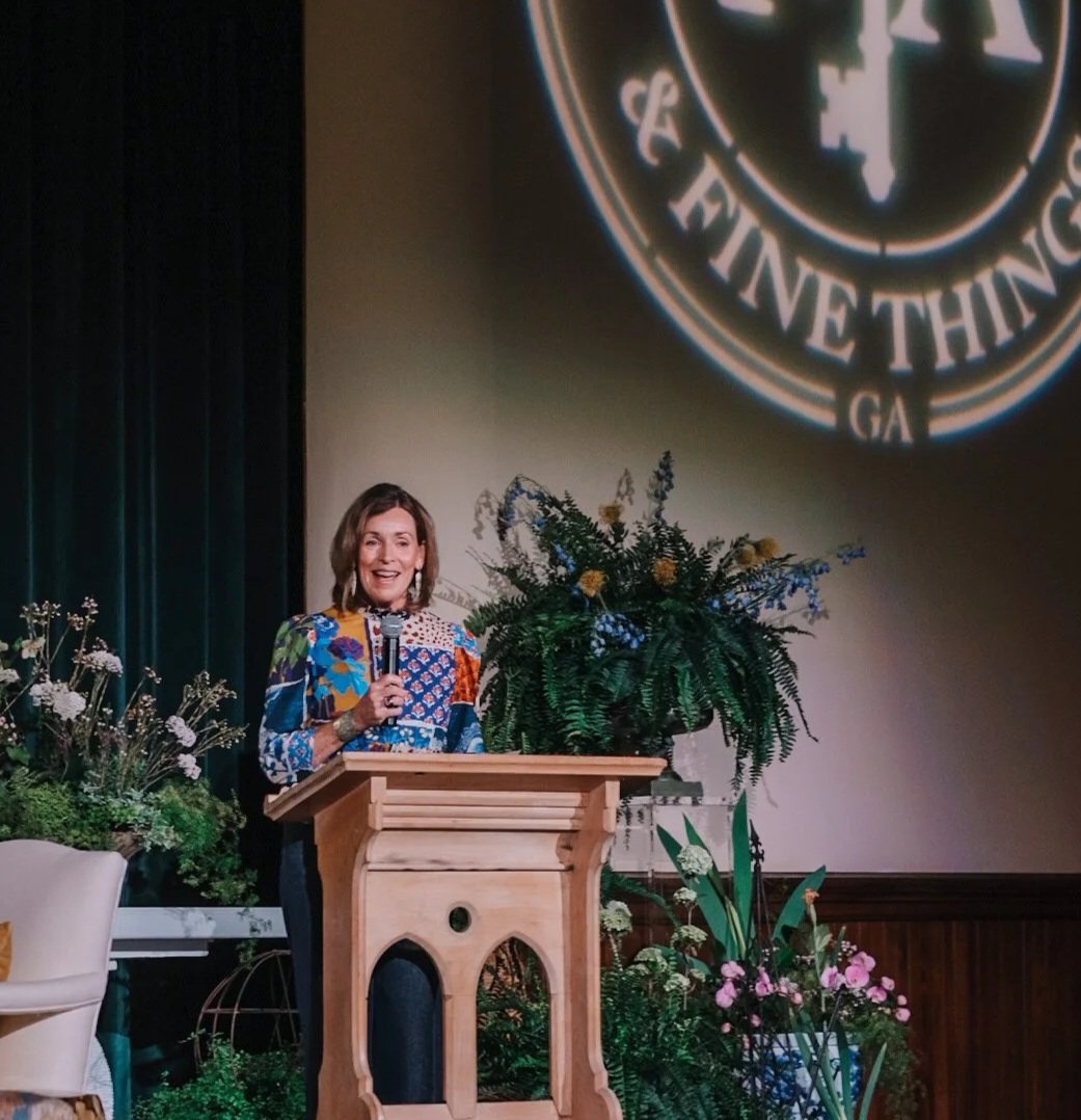 A woman standing at a wooden podium, holding a microphone and smiling, with an arrangement of flowers and plants on the stage. A large projected logo or emblem is partially visible on the wall behind her.