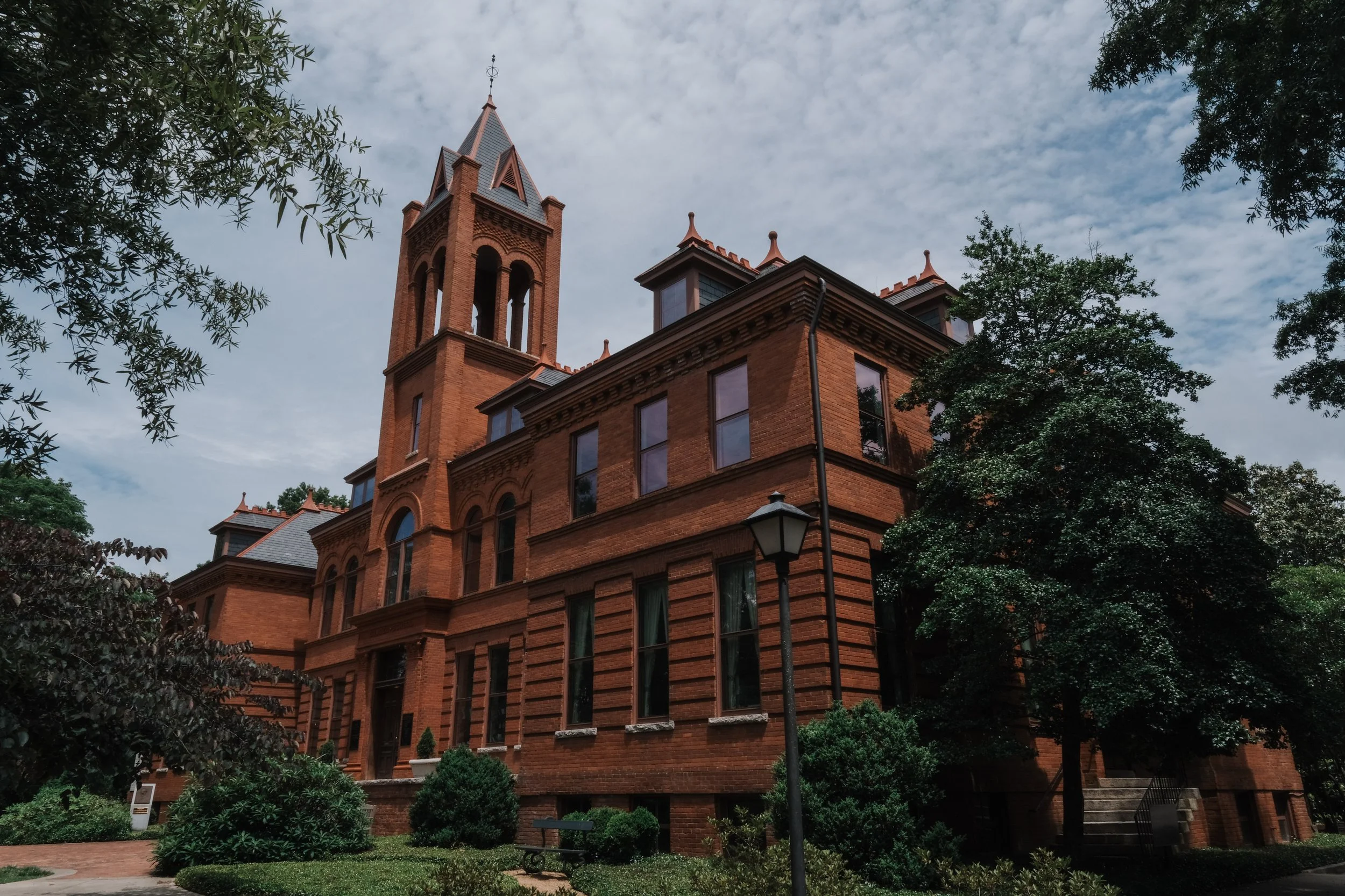 A historic red brick building with a prominent tower and turrets, surrounded by trees, under a cloudy sky.