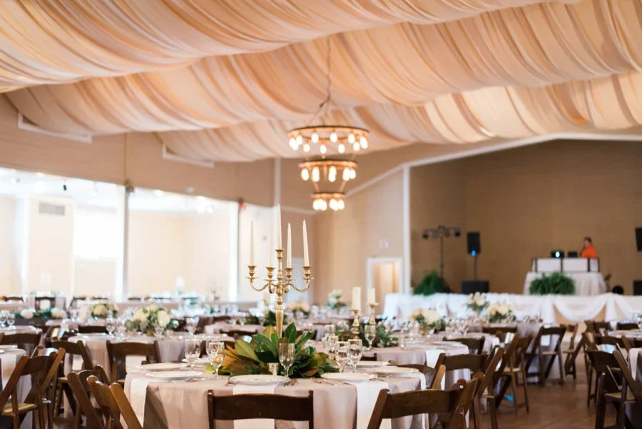 Elegant banquet hall decorated for a wedding reception or special event with round tables, floral centerpieces, candelabras, and a DJ or dance area in the background.
