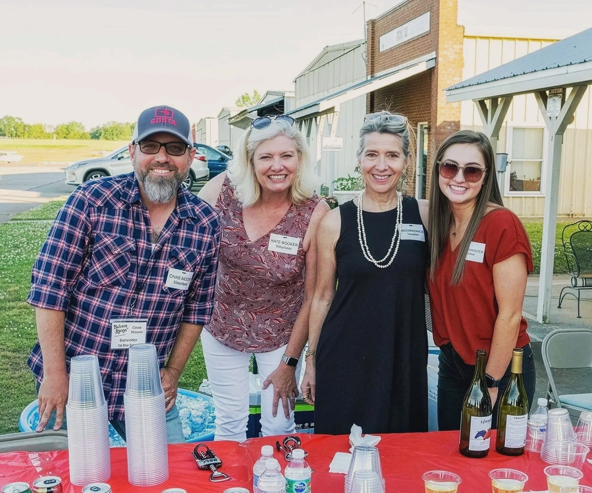 Four smiling people standing outdoors, two women and two men, behind a table with cups, bottles, and snacks, in front of a building with a parking lot and cars in the background. They are wearing name tags, suggesting a social or community event.