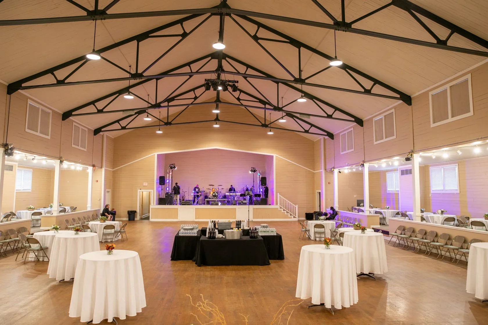 Large indoor event space with a stage at the far end, set up for a live band, with high ceilings and exposed beams. Round tables with white tablecloths and small floral centerpieces are arranged around the room, with chairs placed around each table. The room has soft lighting, creating a festive atmosphere.