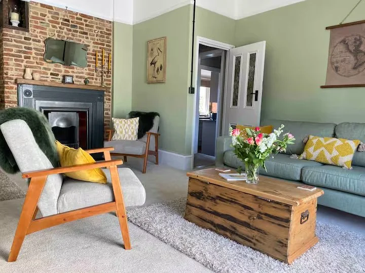 Living room with green walls, fireplace, armchair with yellow pillow, wooden chair, sofa with yellow pillows, wooden coffee table with a flower vase, artwork, and open door leading to another room.