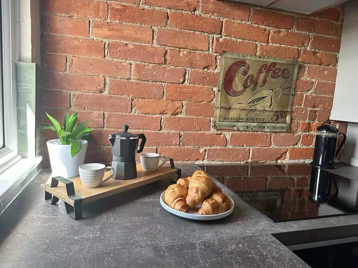 Breakfast time! Kitchen countertop with a small coffee pot, two cups, a plant in a white pot, and a plate of croissants near a brick wall with a vintage coffee sign.