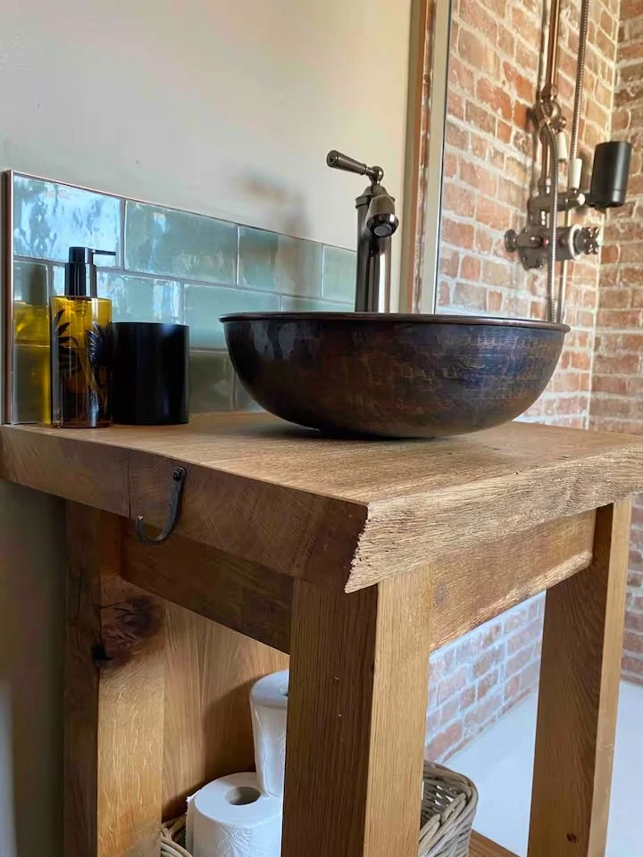 Close-up of a rustic wooden bathroom vanity with a dark wooden vessel sink and chrome faucet, soap dispensers, and a soap container. The background features an exposed brick wall and part of a mirror.
