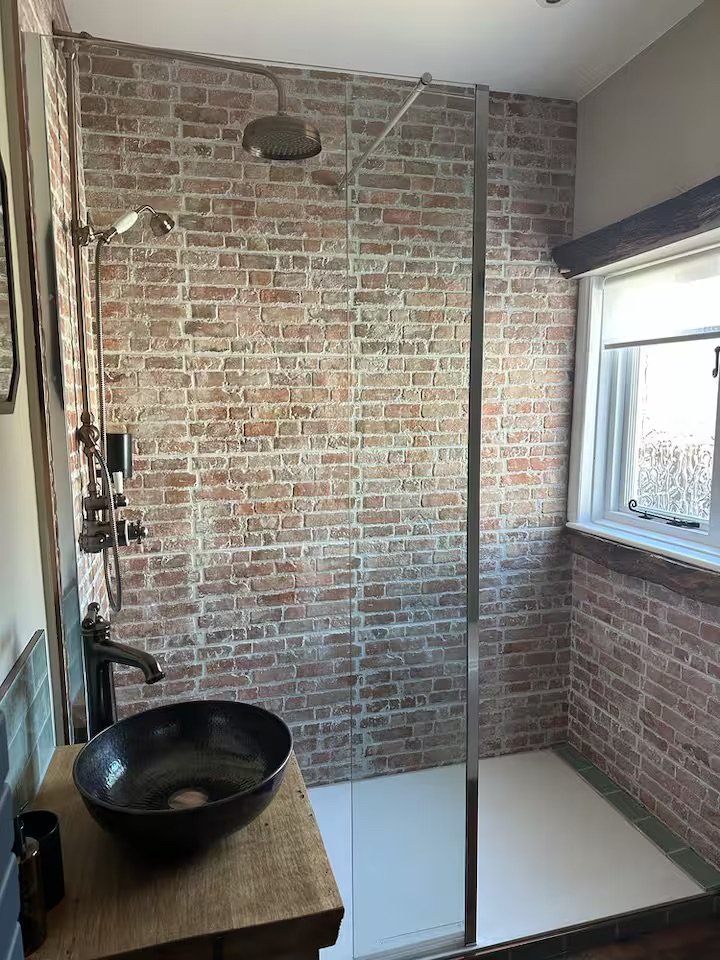 Contemporary bathroom with exposed brick wall, walk-in shower with clear glass door, black vessel sink on wooden vanity, black fixtures, and a window with blinds.