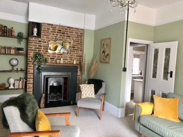 Living room with a brick fireplace and green walls, decorated with plants, shelves, and artwork. There are armchairs with yellow and gray pillows, a gray sofa, and a doorway leading to another room.