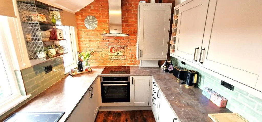 Modern kitchen with gray cabinets, dark marbled countertops, green backsplash tiles, exposed brick wall, built-in oven, small appliances, and decorative plants.