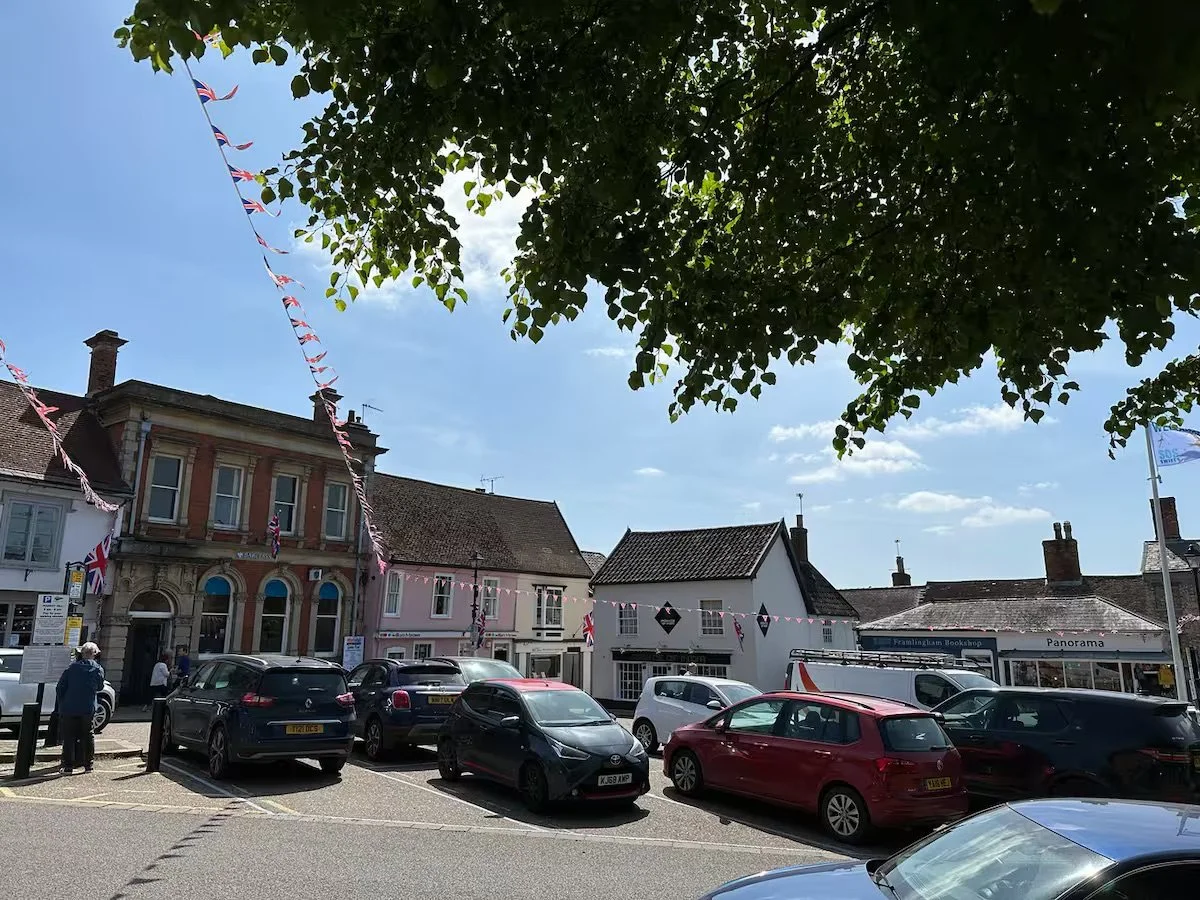 Street scene in the small town of Framlingham decorated with celebrating flags, with parked cars, houses, and trees under a partly cloudy sky.