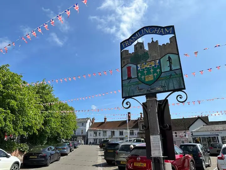 Street scene in Framlingham with parade flags, cars parked along the street, a sign displaying the town's coat of arms, and trees under a cloudy sky.