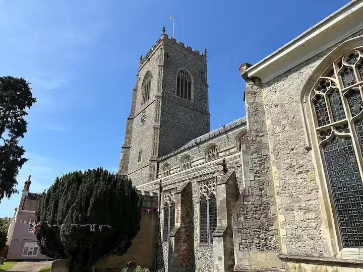A tall historic stone church tower with arched windows, adjacent to a stone church building with stained glass windows, under a clear blue sky.