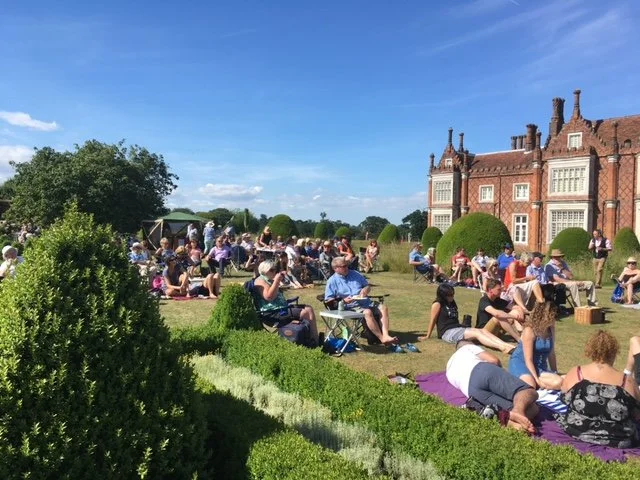 People sitting and lying on the grass at a garden during daytime, with a historic brick building in the background.