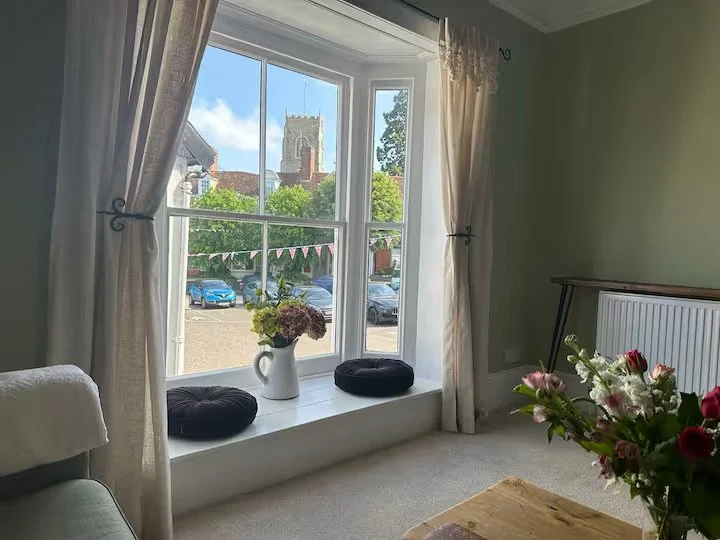 Interior of the cozy living room with a large window, beige curtains, a white windowsill with a vase of flowers, two black cushions, and a view of a church with a tower outside.