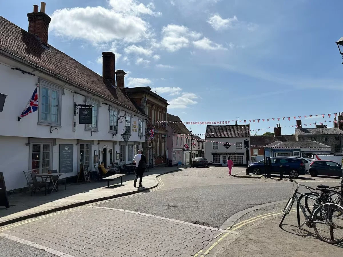 A picture of Framlingham in the UK with old buildings, hanging bunting flags, and parked cars on a sunny day. Some people are walking and sitting outside.