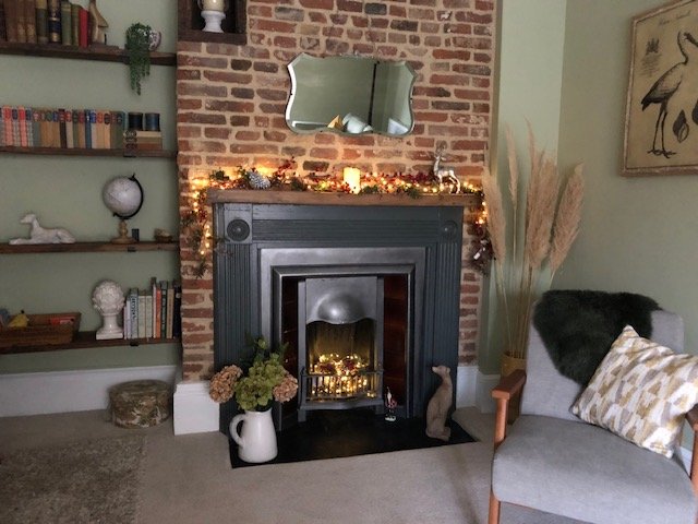 Living room with a brick fireplace decorated with string lights and candles, a mirror above the mantel, a bookshelf with books and decor, a potted plant, and a gray armchair with a patterned pillow. The living room with the fireplace invites to relax
