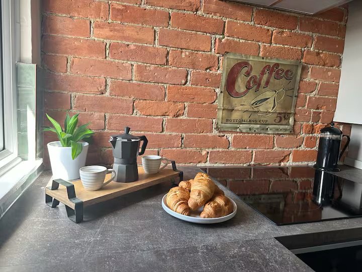 Breakfast time! Kitchen countertop with a small coffee pot, two cups, a plant in a white pot, and a plate of croissants near a brick wall with a vintage coffee sign.