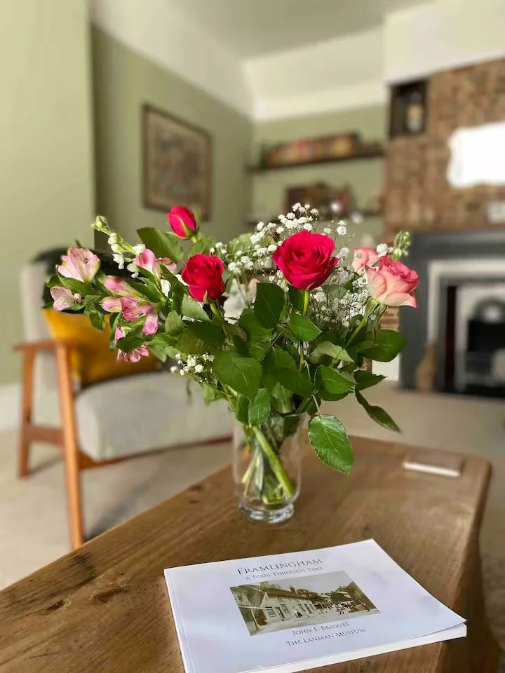 A bouquet of pink and red roses with white baby's breath in a clear glass vase on a wooden table, with a book titled "FRAILTINGHAM" in the foreground, in a living room with green walls, a beige couch, and a brick fireplace.