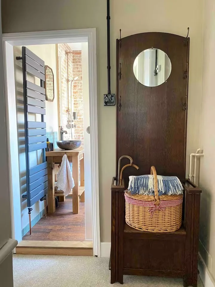 A vintage wooden washstand with a built-in wicker laundry basket and a striped cloth cover, positioned against a wall in a room. To the left, a doorway shows a glimpse of a bathroom with a modern sink, wooden vanity, and a black towel warmer on the w