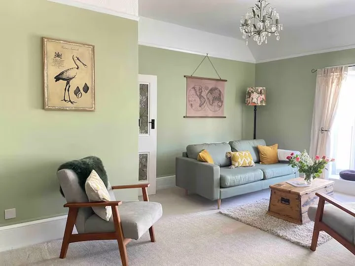 Living room with light green walls, a gray sofa with yellow and white pillows, a wooden coffee table with flowers, two armchairs, a chandelier, and wall art.