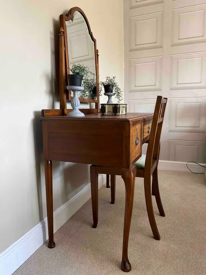 A vintage wooden vanity table with a mirror, decorated with two potted plants, a decorative box, and a wooden chair in a room with beige carpet and paneled white walls.