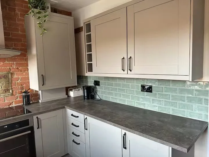Kitchen with beige cabinets, gray countertop, green brick-style tile backsplash, and an exposed brick wall on the left, featuring a coffee maker and other appliances.