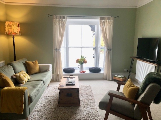 Bright living room with pale green walls, a large window with white curtains, a cream sofa with yellow and patterned pillows, a wooden coffee table, a gray armchair with yellow accent pillow, a TV on a black stand, and a floor lamp with a floral lamp