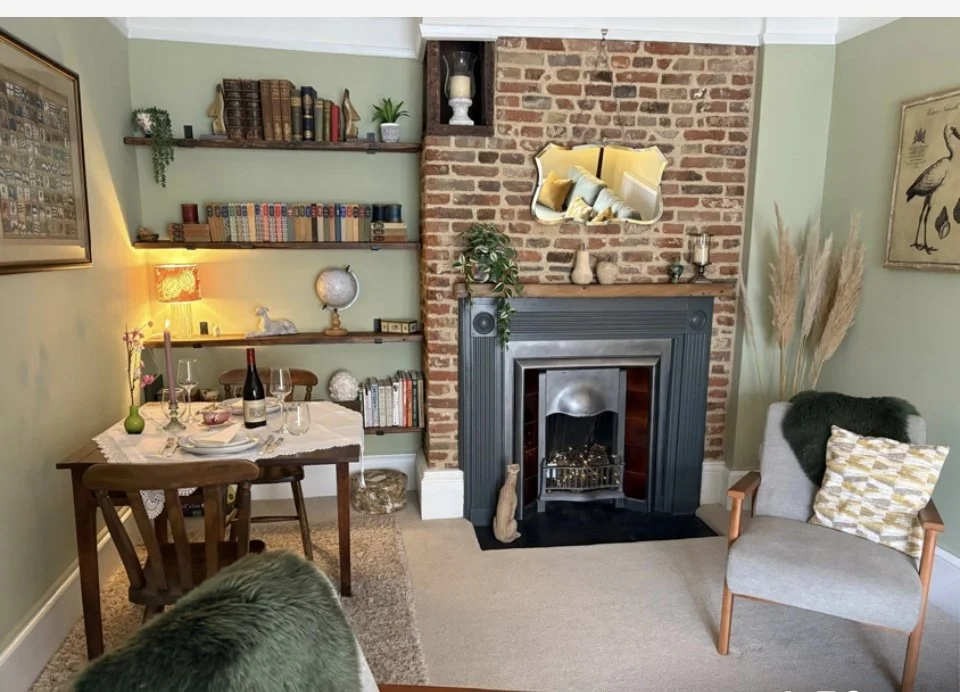 Cozy living room with brick fireplace, green walls, a wooden dining table set for dinner, bookshelves filled with books, and decorative plants and lamps.