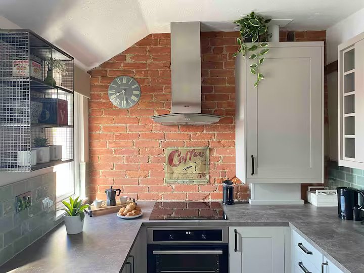 Modern kitchen with exposed brick wall, gray cabinets, stainless steel range hood, black stovetop, coffee sign, and decorative plants.
