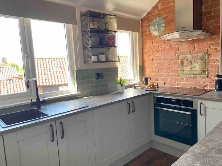 Kitchen with white cabinets, a gray countertop, and a brick accent wall. There is a window above the sink, a wall clock, and a mounted metal shelf with kitchen items. A plant, a coffee maker, and a cutting board with bread are on the counter.