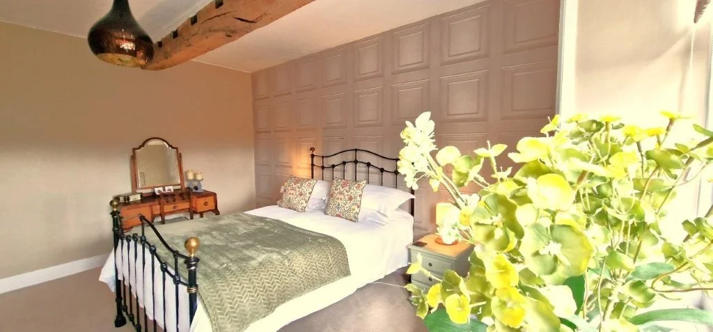 A cozy bedroom with a wrought iron bed, decorative pillows, a green blanket, a wooden vanity with a mirror, a small nightstand, and a large plant near a window with sunlight.