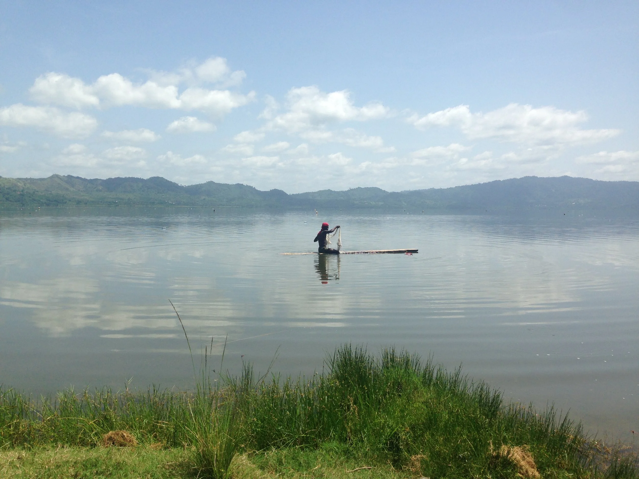 A person sitting on a boat on a calm lake, fishing with a fishing net under a partly cloudy sky, with hills in the background.